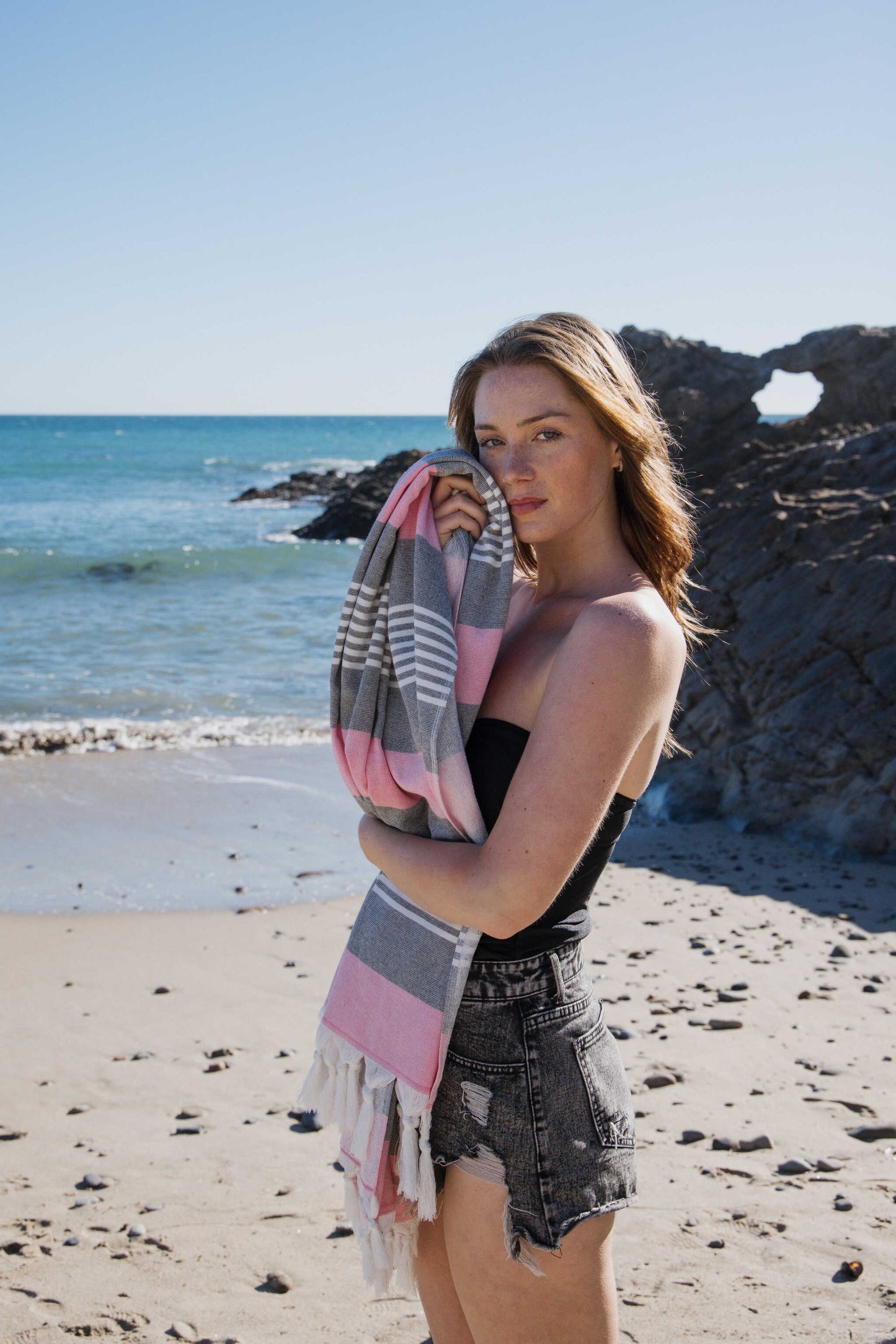Woman holding a striped towel on a beach with ocean and rocks in the background