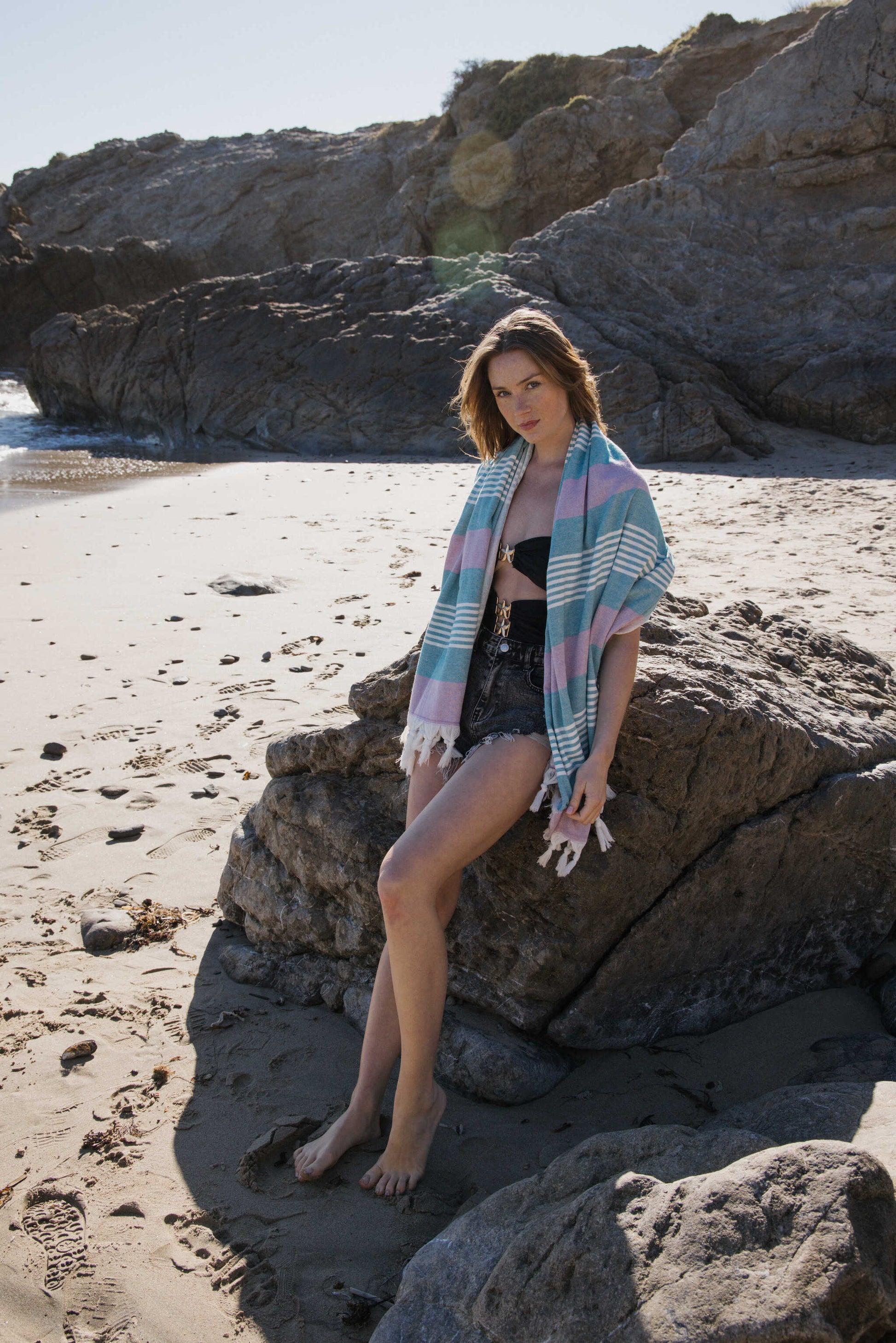 Woman sitting on a rock at the beach with a towel draped over her shoulders.