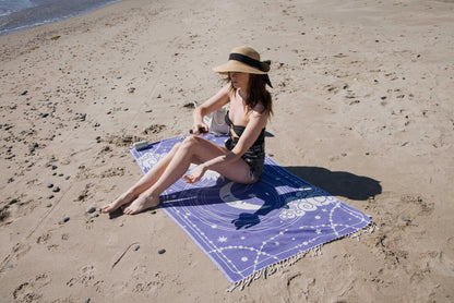 Woman sitting on a purple beach towel with star patterns on a sandy beach.