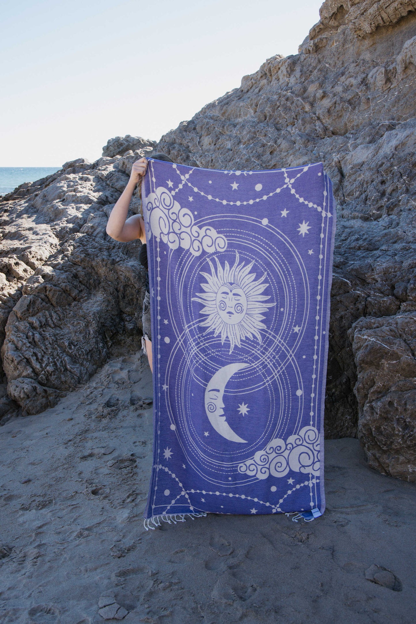 Person holding a purple towel with celestial designs on a rocky beach.