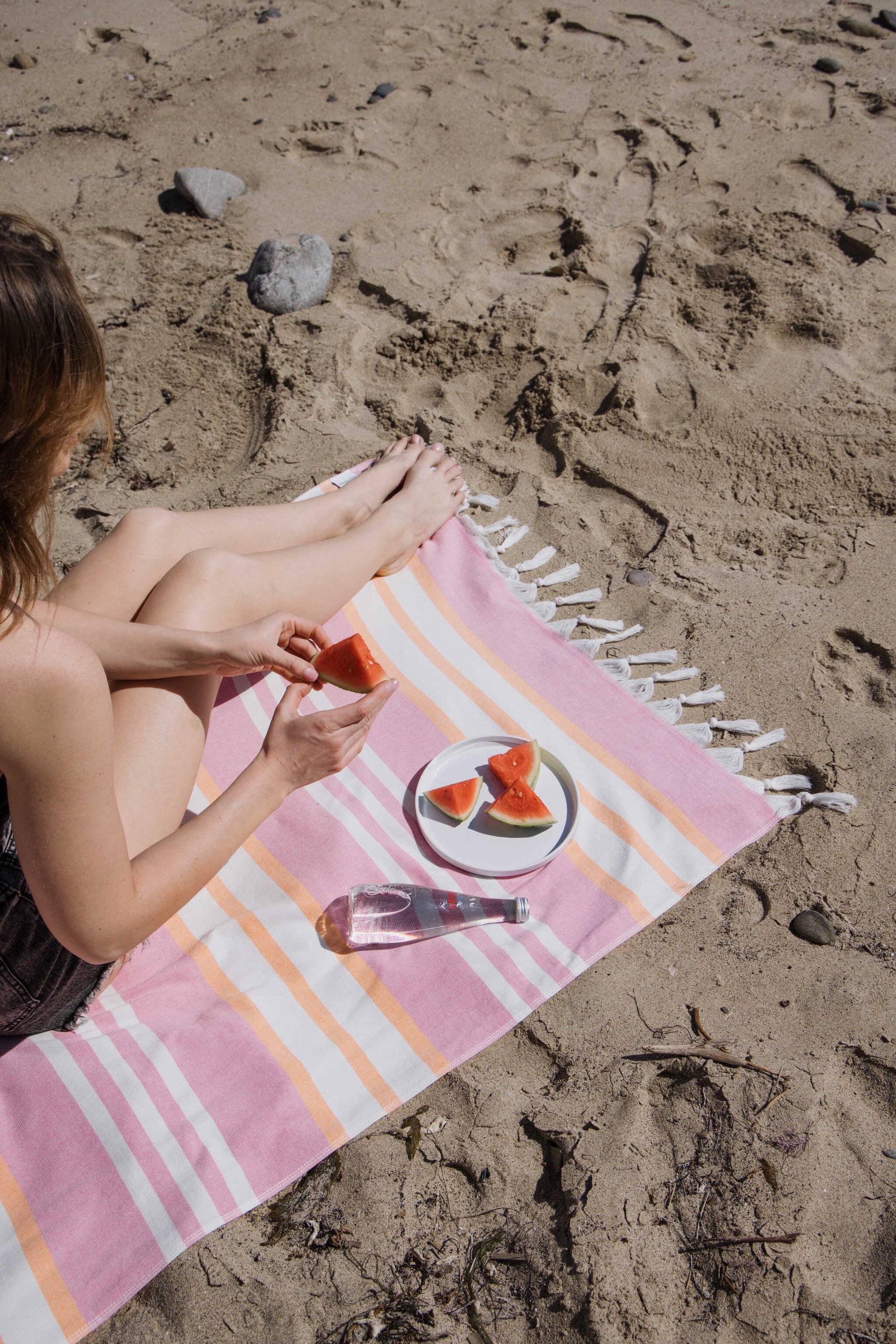 Person sitting on a striped beach towel with watermelon slices on a sandy beach