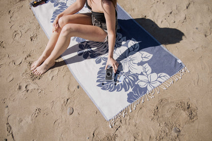 Person sitting on a beach towel with floral patterns, holding a camera on a sandy beach.