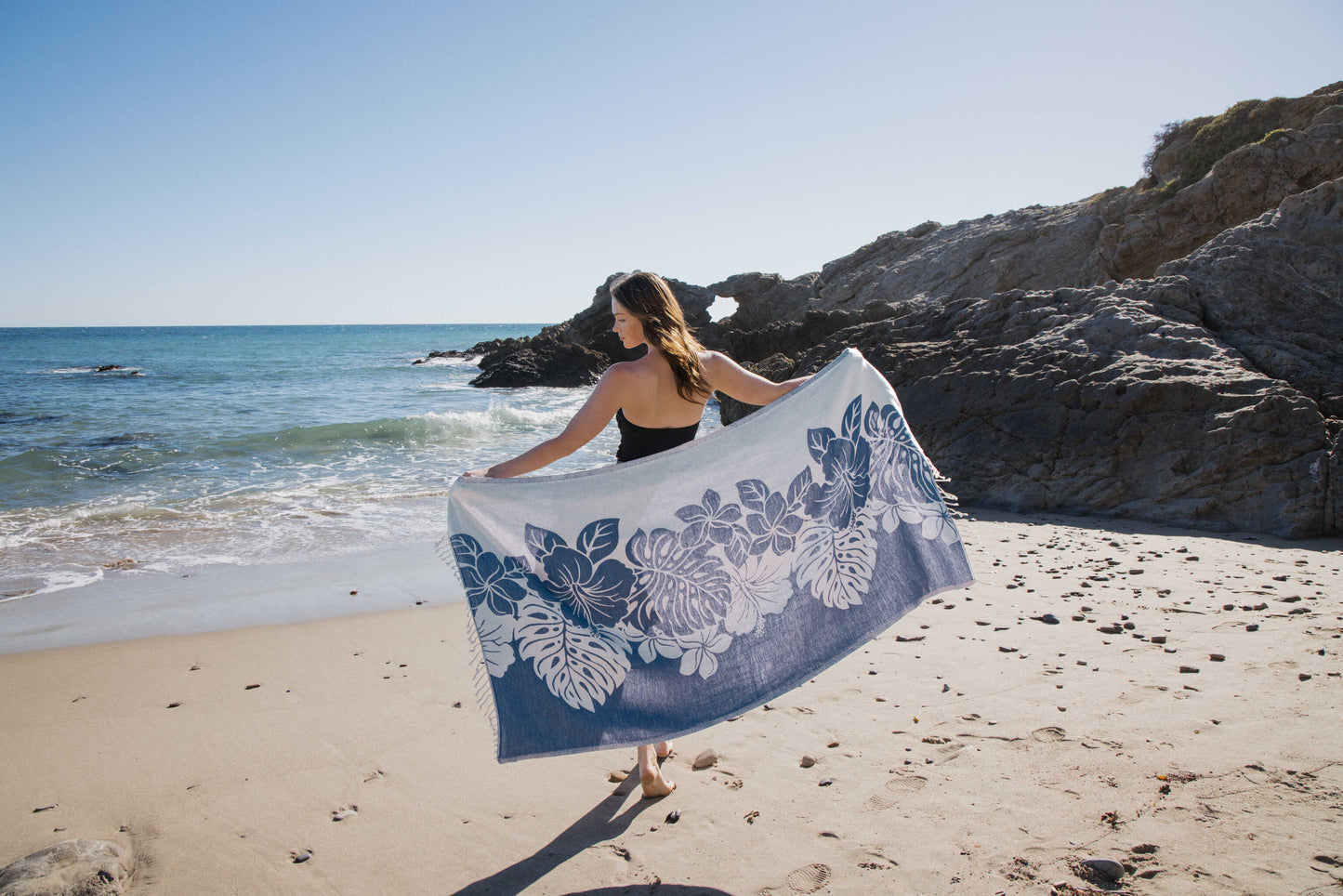 Woman holding a blue floral towel on a sandy beach with ocean and rocks in the background