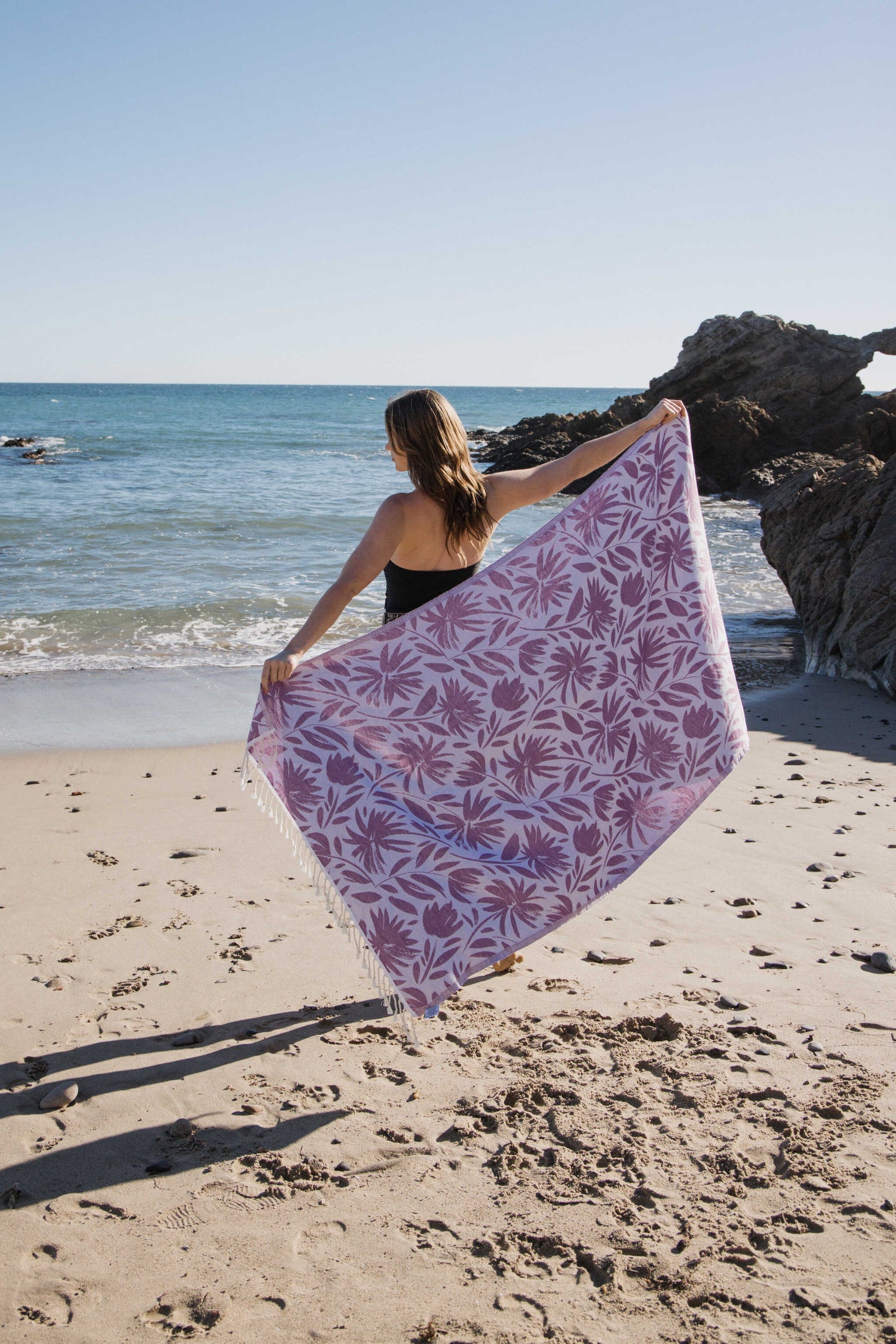 Woman holding a purple floral towel on a beach with ocean and rocks in the background