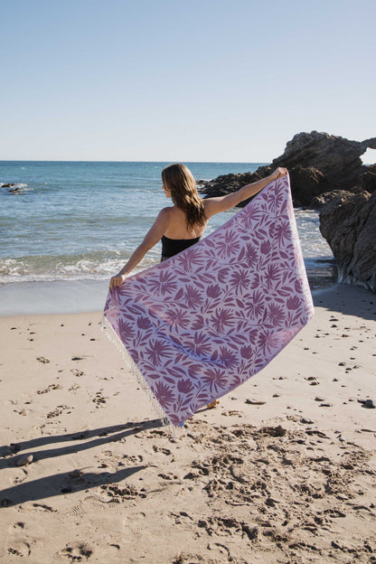 Woman holding a purple floral towel on a beach with ocean and rocks in the background